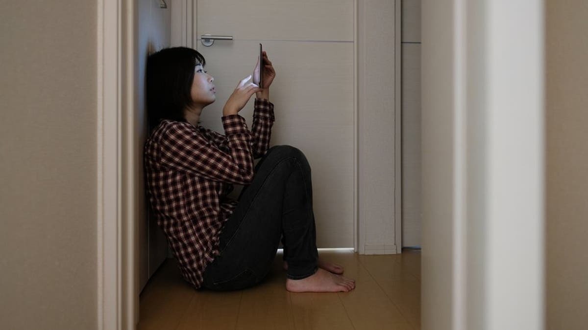 Chica aburrida sentada en el suelo de su casa mirando su celular durante la crisis del hikikomori y la falta de socialización.
